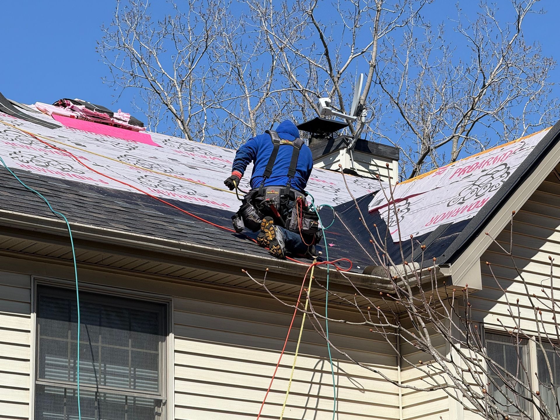 Roofer in blue suit on a roof, attached to safety lines, working on shingles.