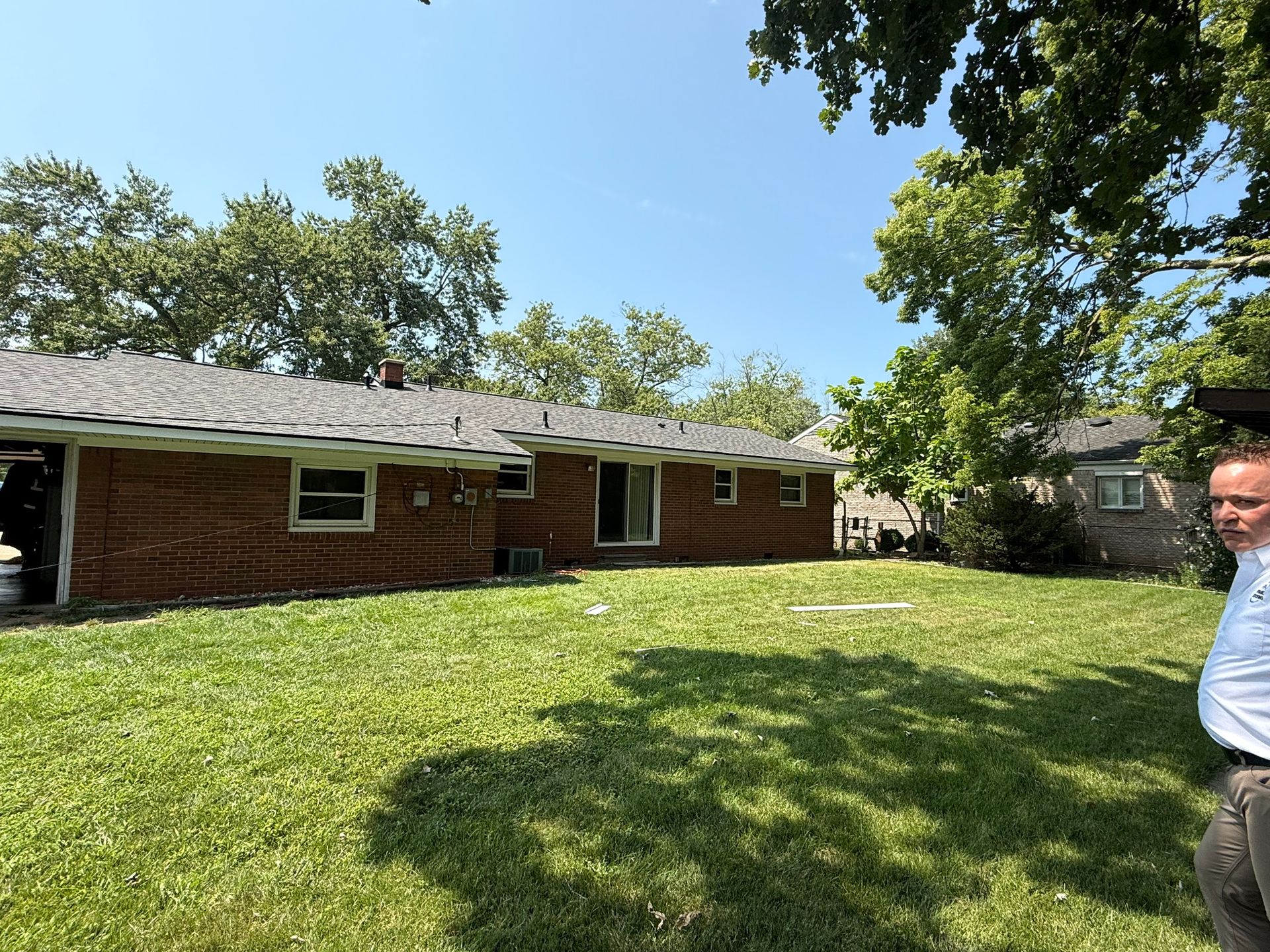 Backyard of a brick house with green grass, trees, and a person in the right corner.
