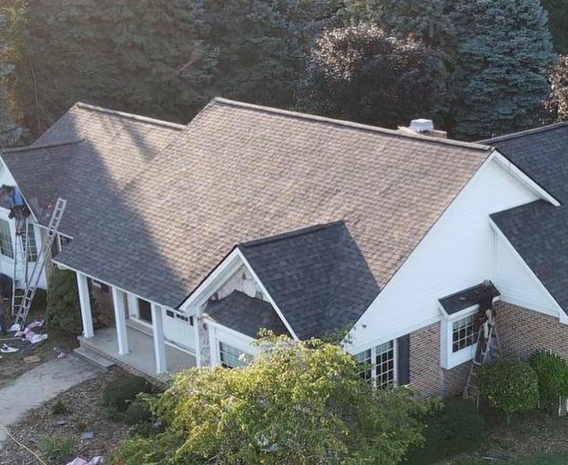 A house with dark roof shingles. Workmen are on a ladder next to the roof. Trees and bushes surround the house.