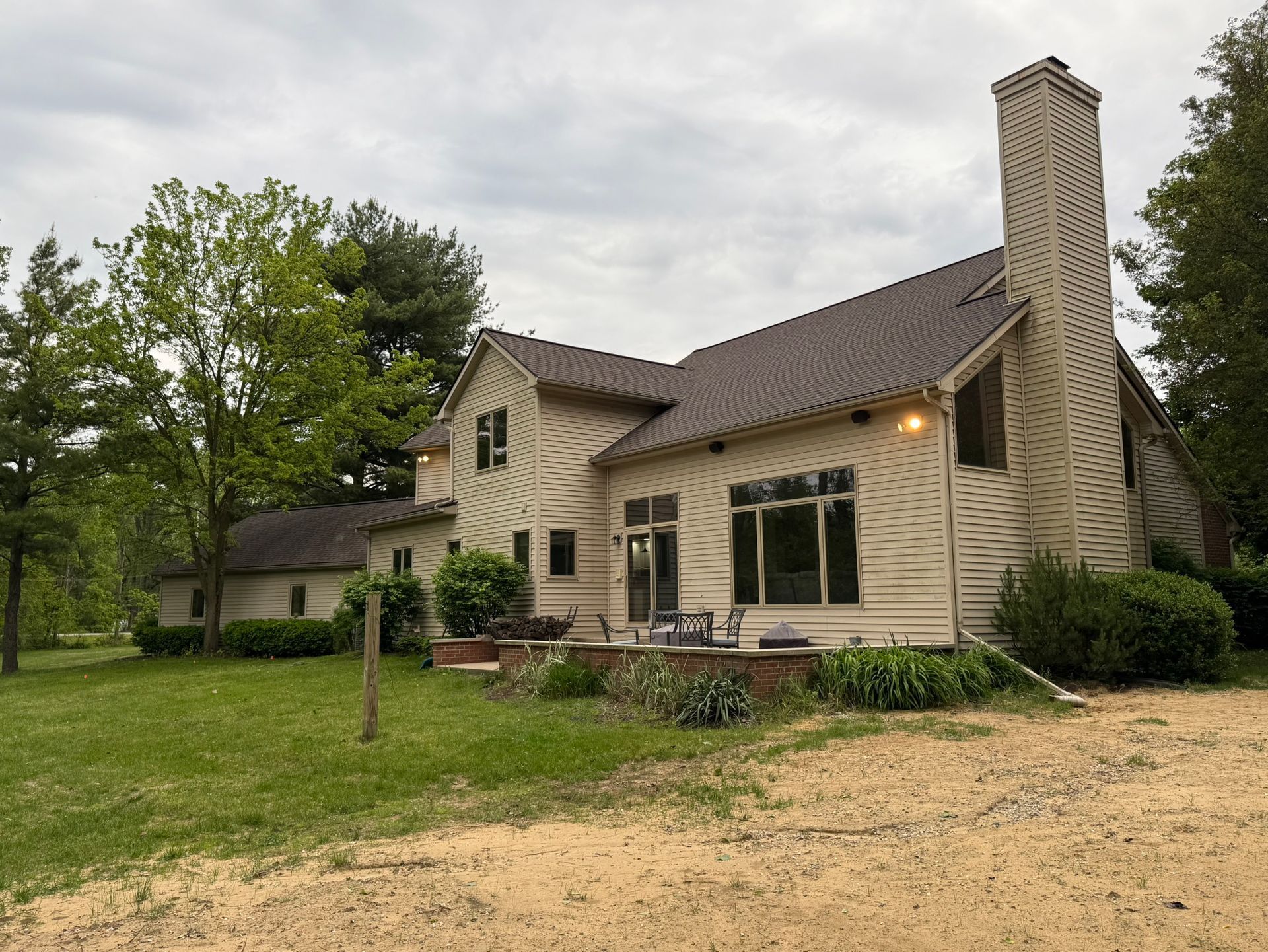 Tan house with brown roof, large windows, and a tall chimney, set on a grassy lawn with trees.