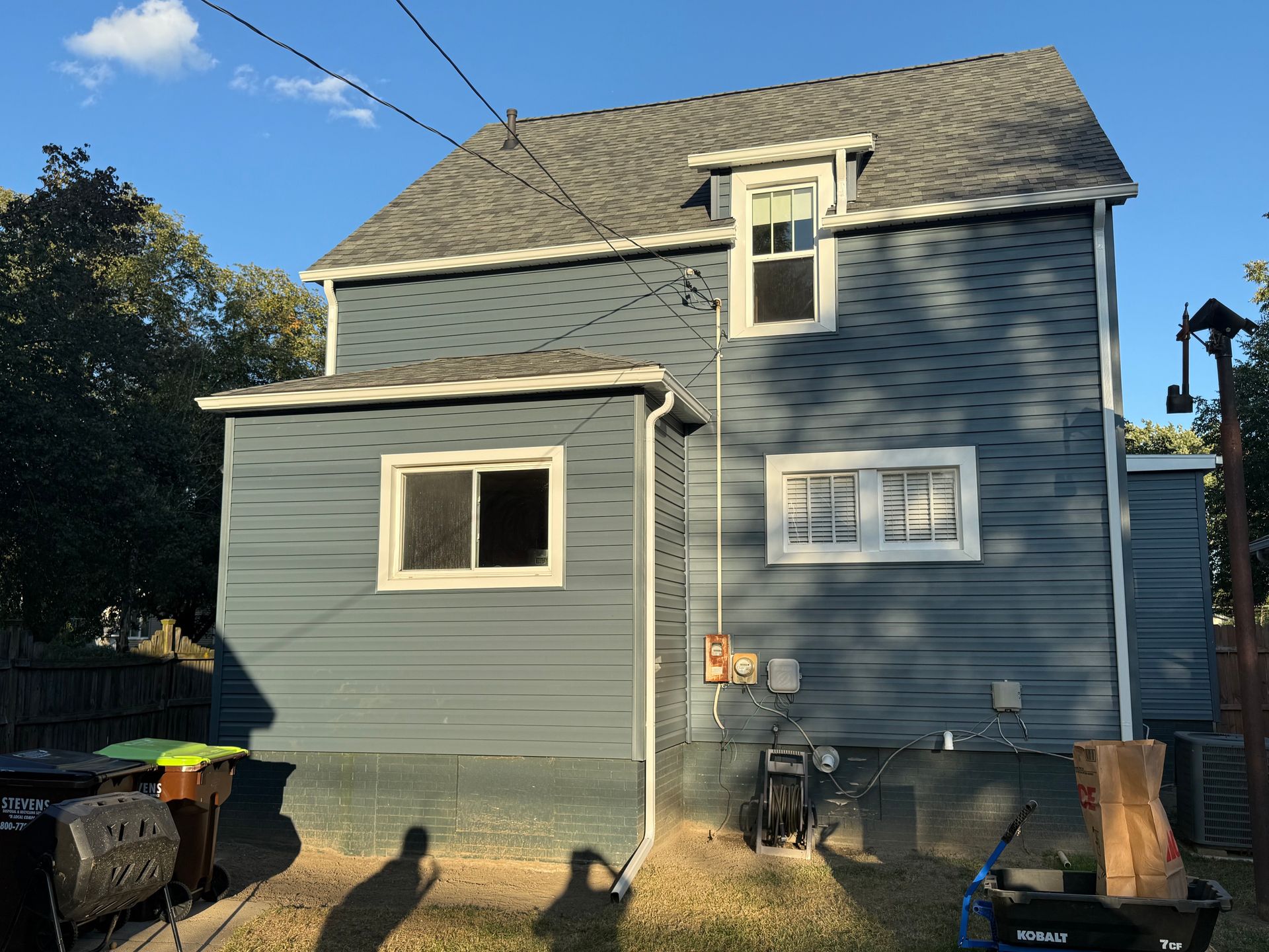 Back of a two-story blue house with white trim, windows, and a gray roof on a sunny day.