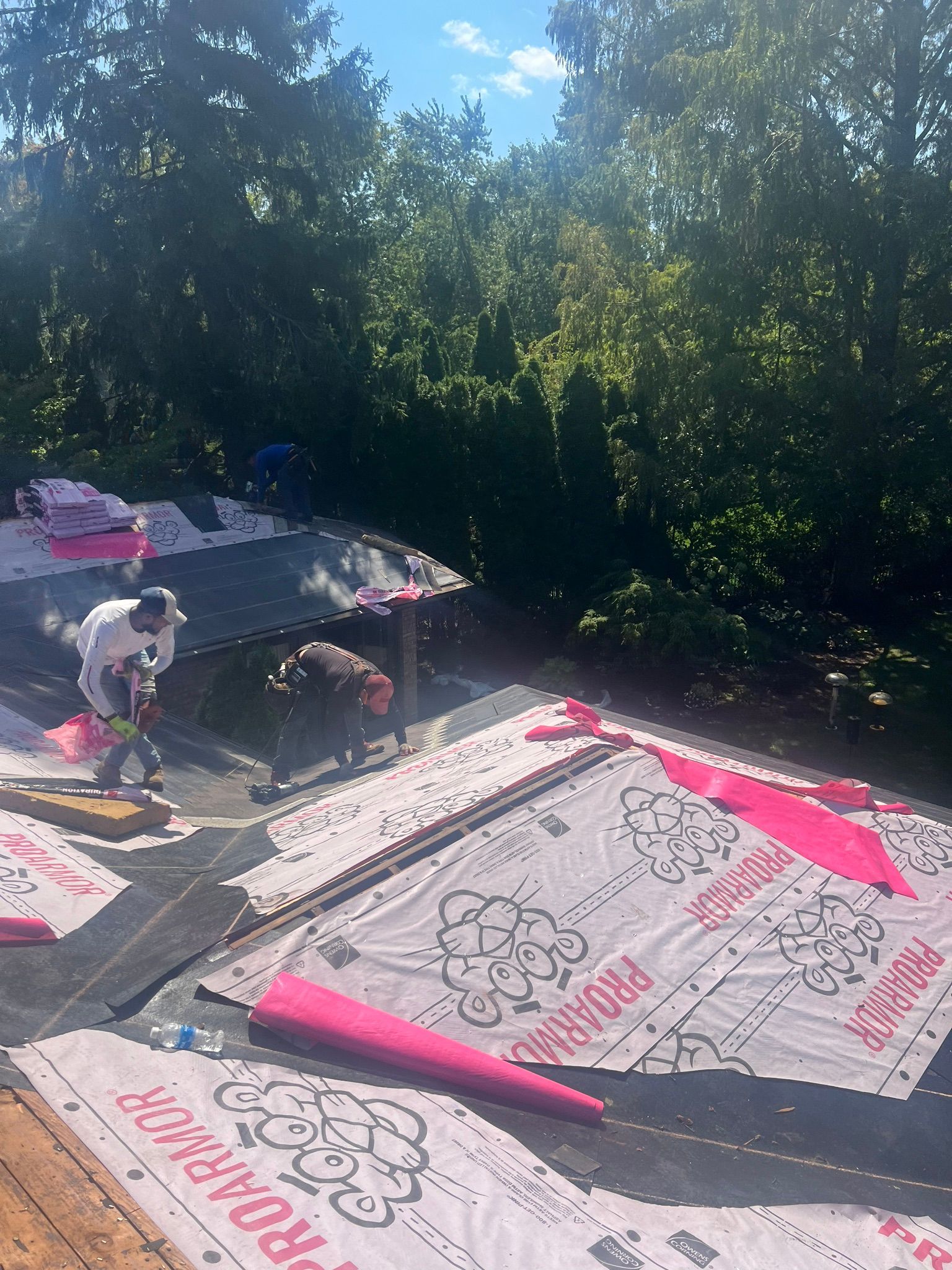 Roofers working on a house roof, using tools, with a forest background and blue sky.