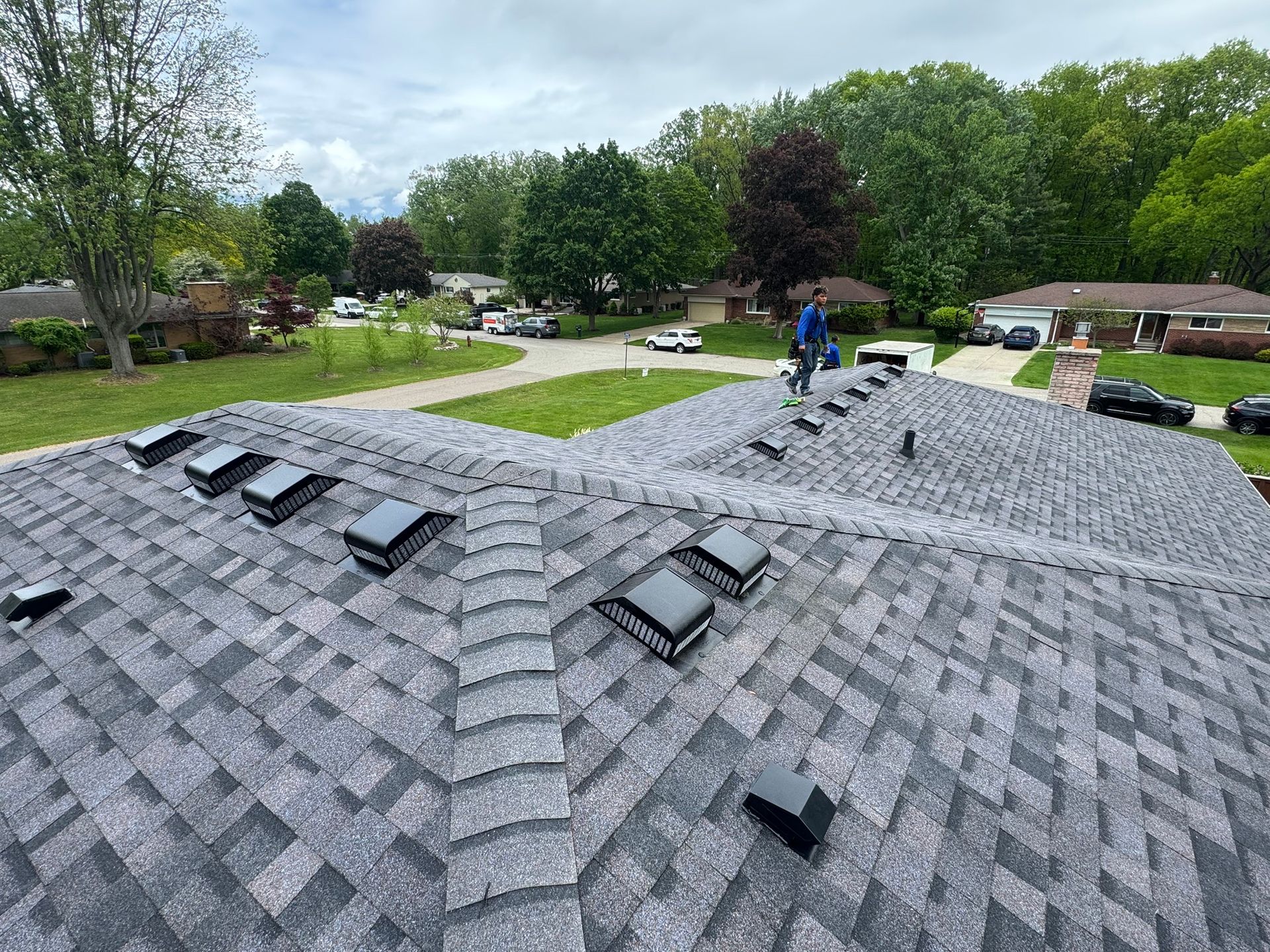 Roofer on a gray shingle roof with vents, working on a house in a residential area with trees.