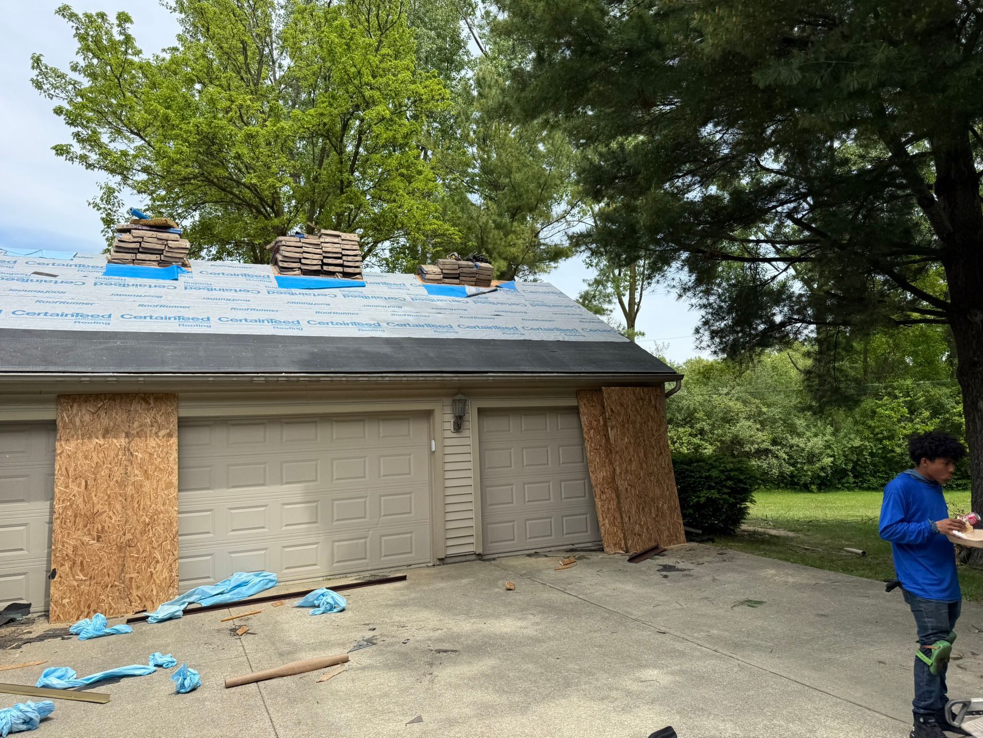 Garage roof under construction; worker, materials, plywood, blue underlayment, shingles stacked.