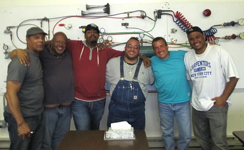 Six men pose in front of a white wall with air brake system diagrams. They are smiling, arms around each other.