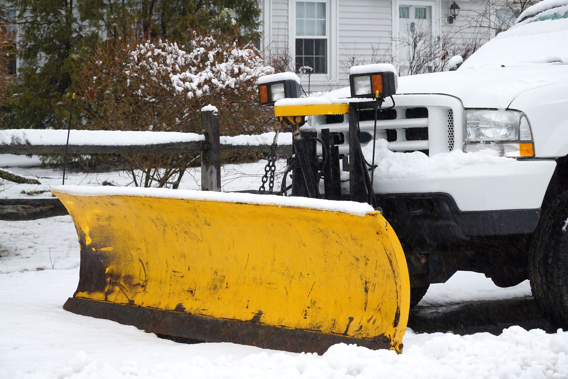 Yellow snowplow attached to a white truck, clearing snow in front of a house.
