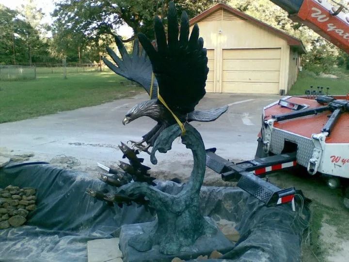 Black bird sculpture beside a garage, partly covered in plastic on a driveway with a trailer nearby