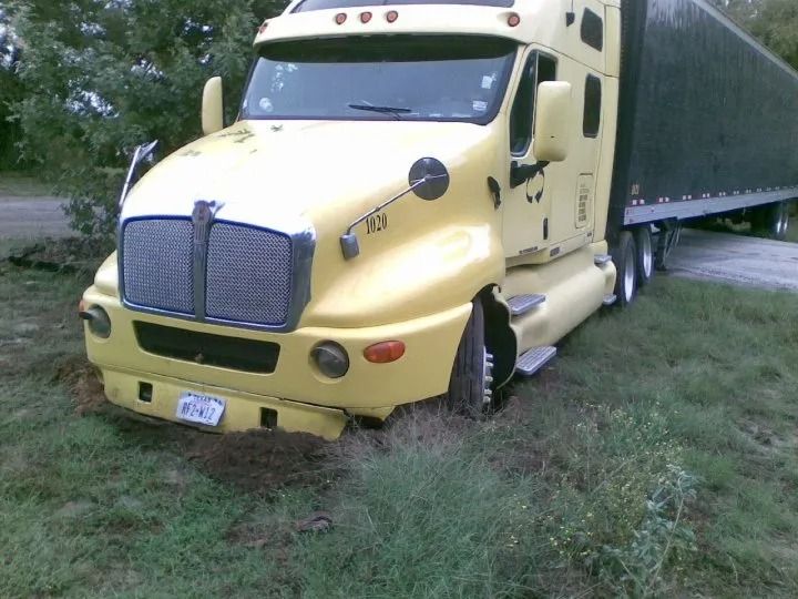 Yellow semi-truck stuck in a grassy ditch, front wheel turned and cab tilted.