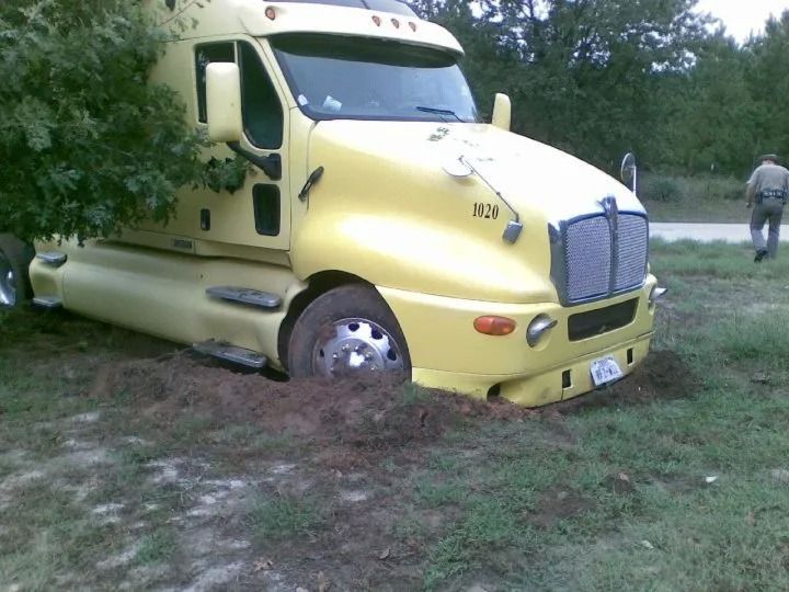 Yellow semi-truck stuck in muddy ground, tilted forward in a grassy field.