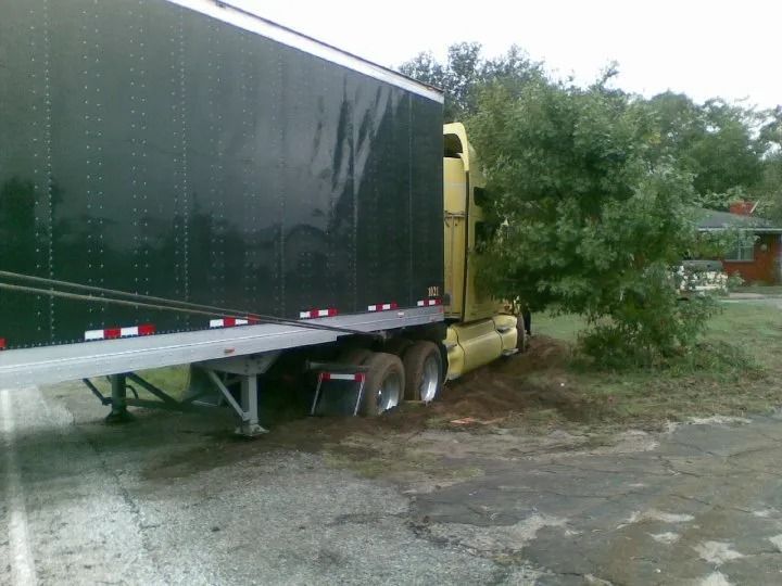 Yellow semi-truck stuck off-road beside bushes on a muddy shoulder
