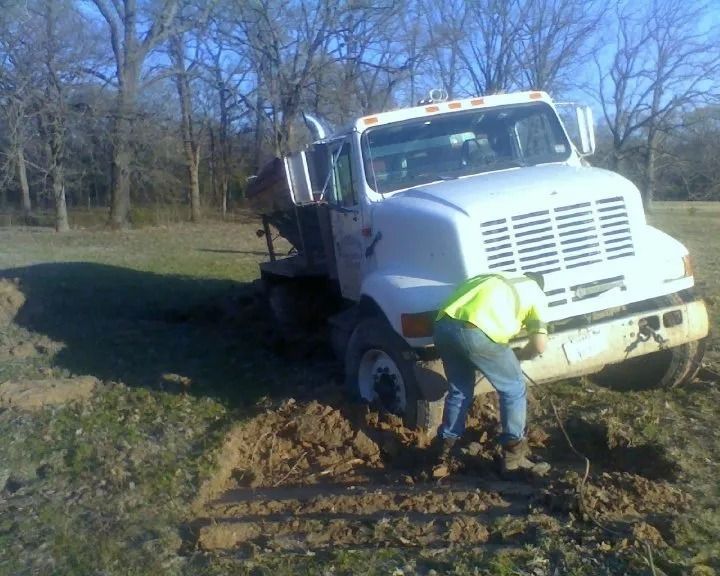 Worker in neon jacket digging near a stuck white truck in a muddy field.