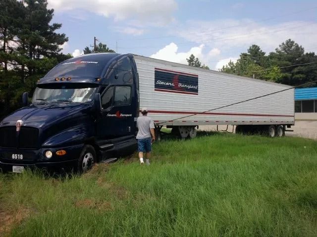 Blue semi-truck with a long white trailer parked in tall grass, with a person standing beside it