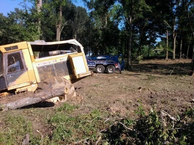 Yellow overturned vehicle in muddy field, with a blue car parked nearby and trees in the background