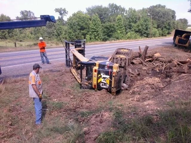 Overturned yellow construction vehicle on a dirt roadside embankment, with two workers nearby.