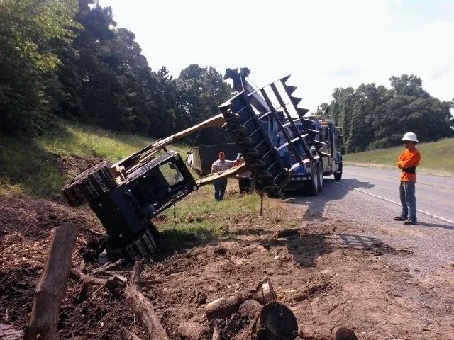 Overturned blue truck on roadside embankment; workers inspect the scene near a gravel road.