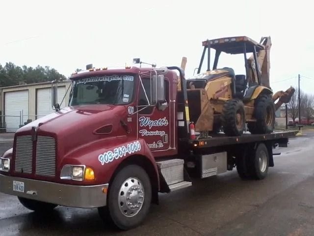 Red tow truck carrying a yellow backhoe on a flatbed, parked outdoors.