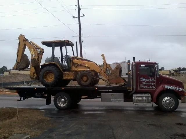 Yellow backhoe loader on a flatbed truck in a rainy parking lot