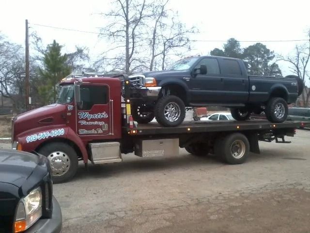 Red tow truck carrying a black pickup on a flatbed in a dirt lot