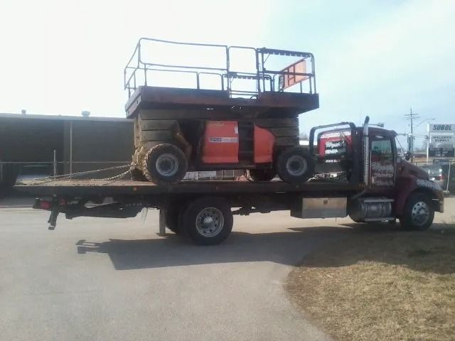 Flatbed tow truck carrying a small red boom lift on a roadside under a bright sky