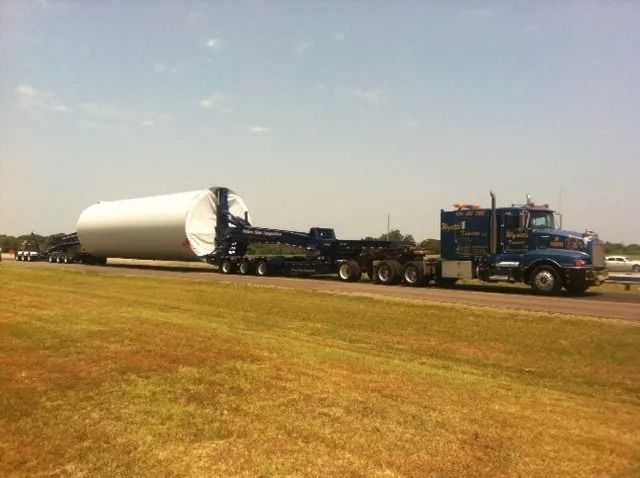 Blue semi-truck hauling a large white cylindrical load across a grassy field under a clear sky