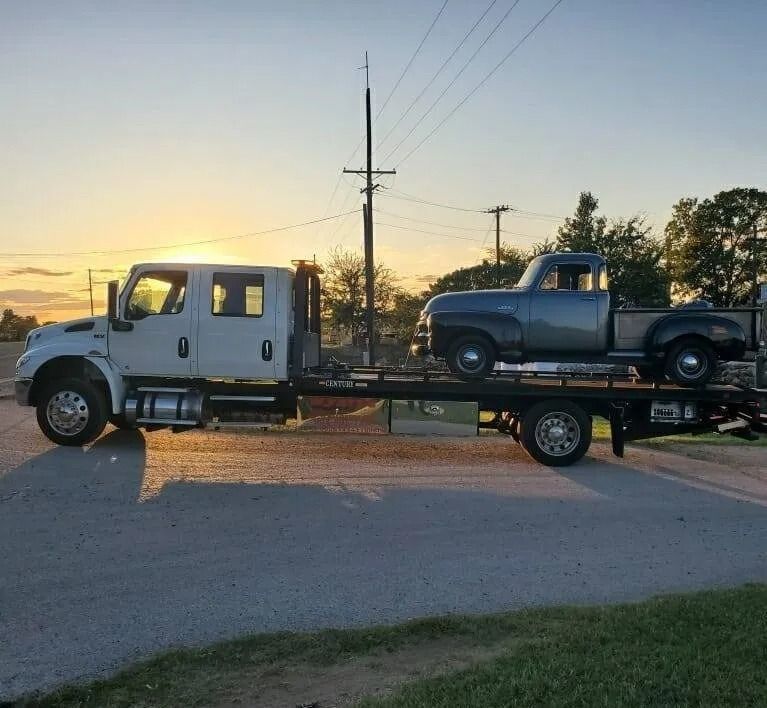 Flatbed tow truck carrying a vintage blue pickup at sunset on a quiet road