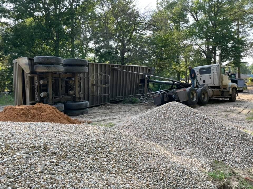 Dump truck overturned on gravel road beside a loader, with piles of dirt and gravel under trees