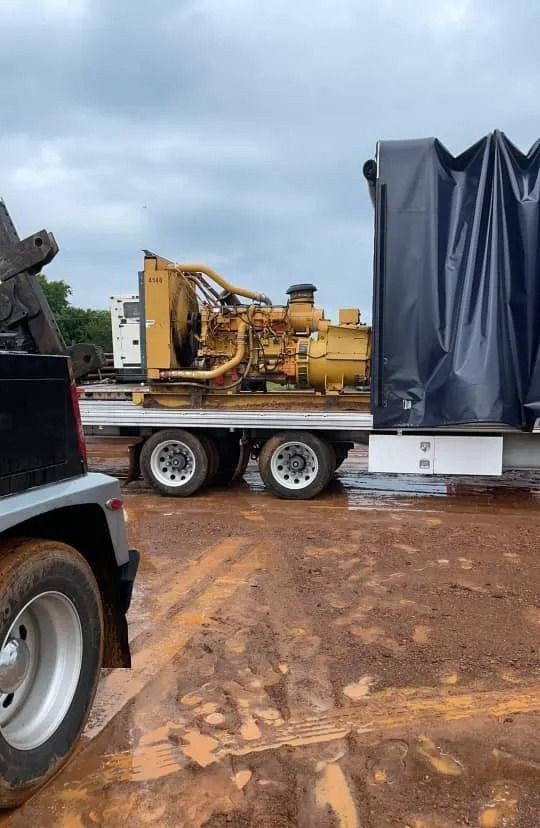 Yellow industrial machine on a trailer beside a covered load in a muddy lot under cloudy skies