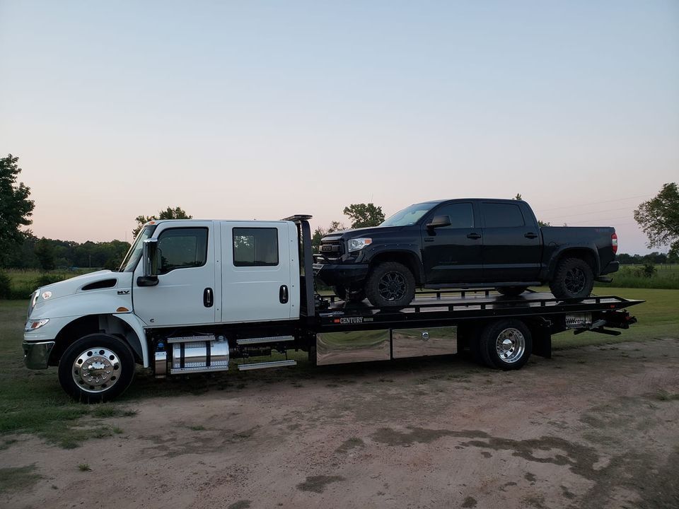 White and black pickup trucks on a flatbed trailer in a dirt lot at dusk