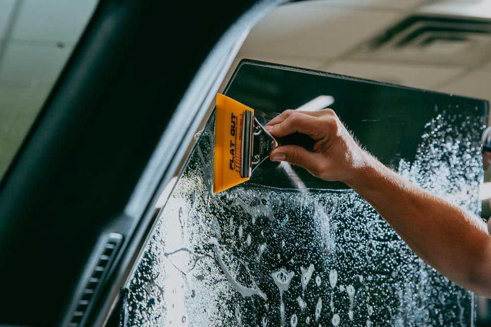Person applying tint to a car window with a squeegee in a shop.