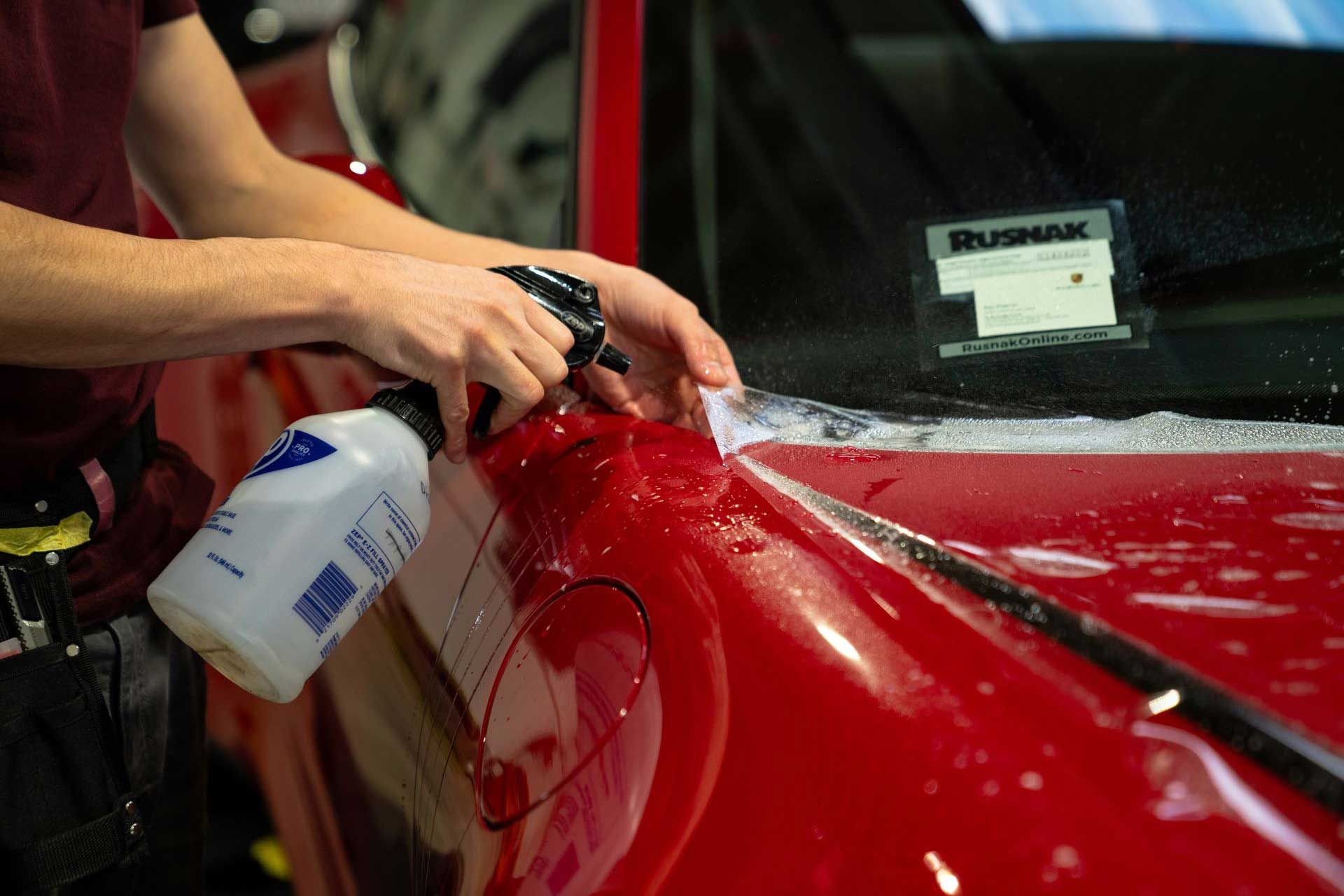 Person spraying a red car with liquid while applying a protective film.