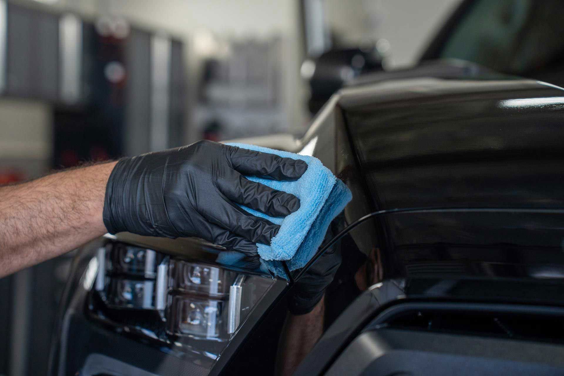 Gloved hand polishing black car with a blue cloth, in a garage.