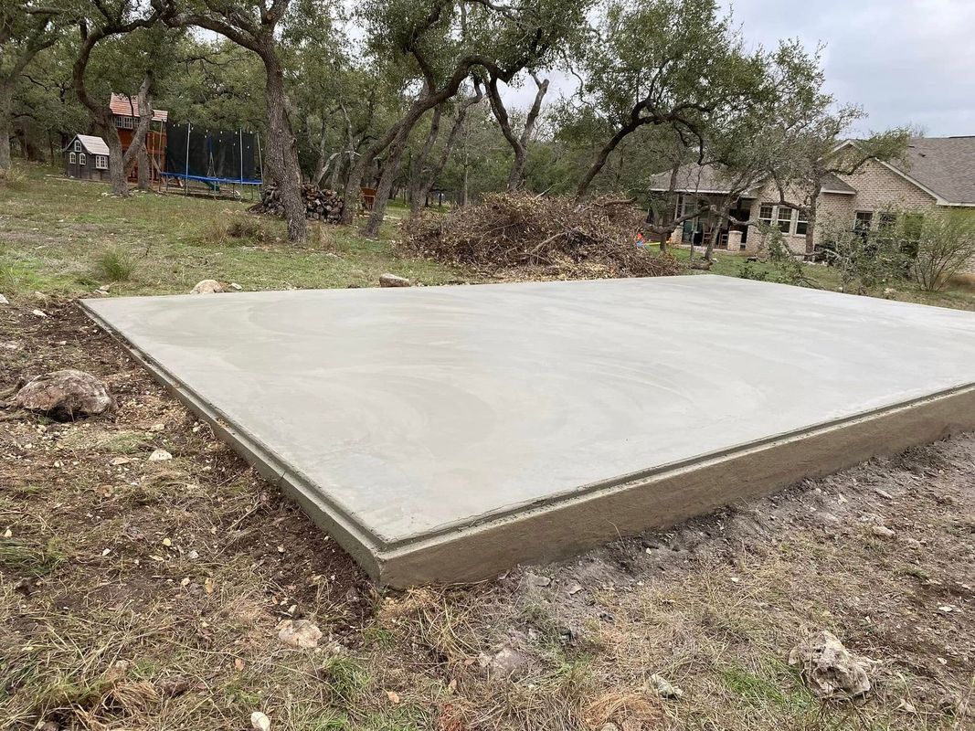 A freshly poured concrete slab, outdoors on a slightly sloped, grassy lot.