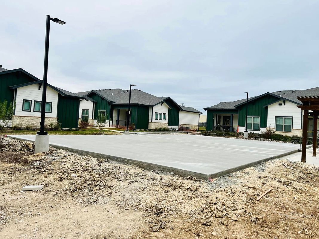 Concrete patio under construction, with green and white houses in the background. Overcast sky.