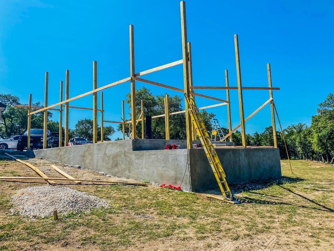 Concrete foundation with wooden beams and posts under construction on a grassy lot under a blue sky.