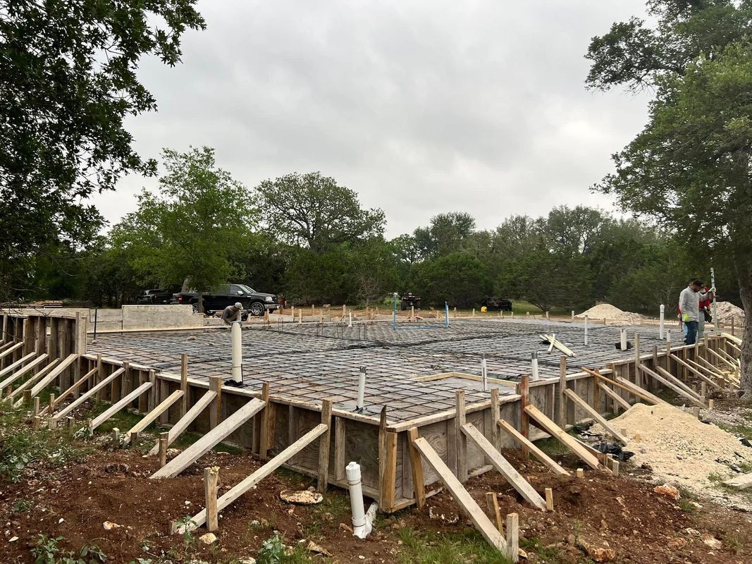 Foundation of a house under construction, with wooden supports and plumbing pipes visible in a wooded area.
