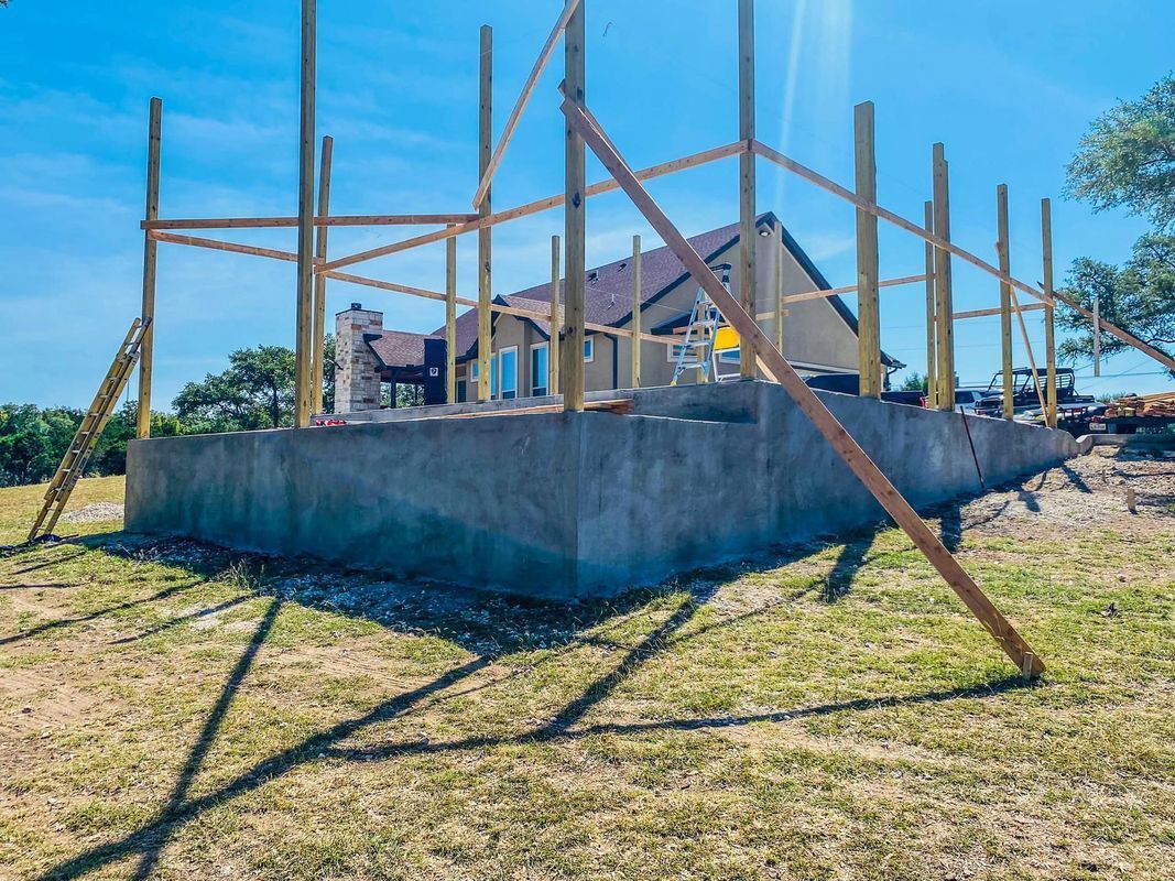Construction site: concrete foundation with wooden posts and framing, house in background, sunny outdoors.