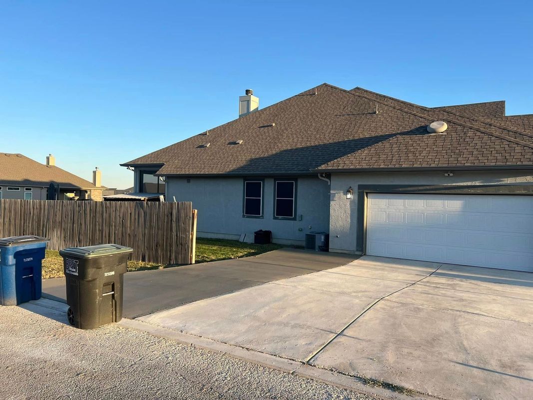 Back of a light blue house with a gray roof, driveway, fence, and trash cans, under a clear blue sky.