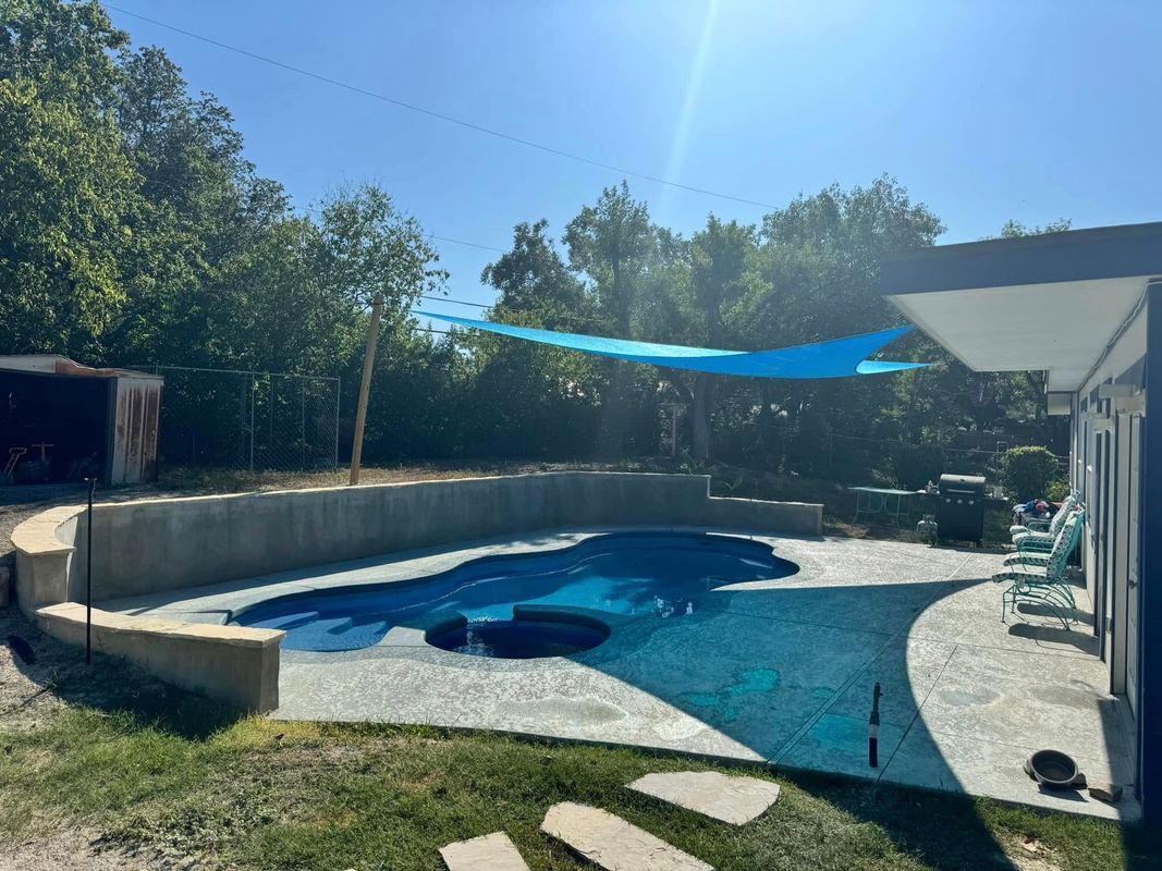 Blue pool with a shade sail, surrounded by concrete and lawn on a sunny day.