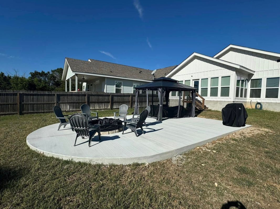 Concrete patio with fire pit and gazebo, surrounded by chairs, grass, and houses on a sunny day.