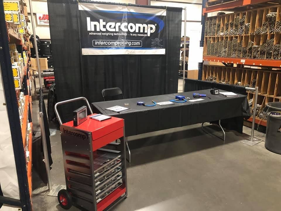 Intercomp booth with black backdrop, table displaying products, and a red tool cart. Display is in an industrial setting with shelves of parts.