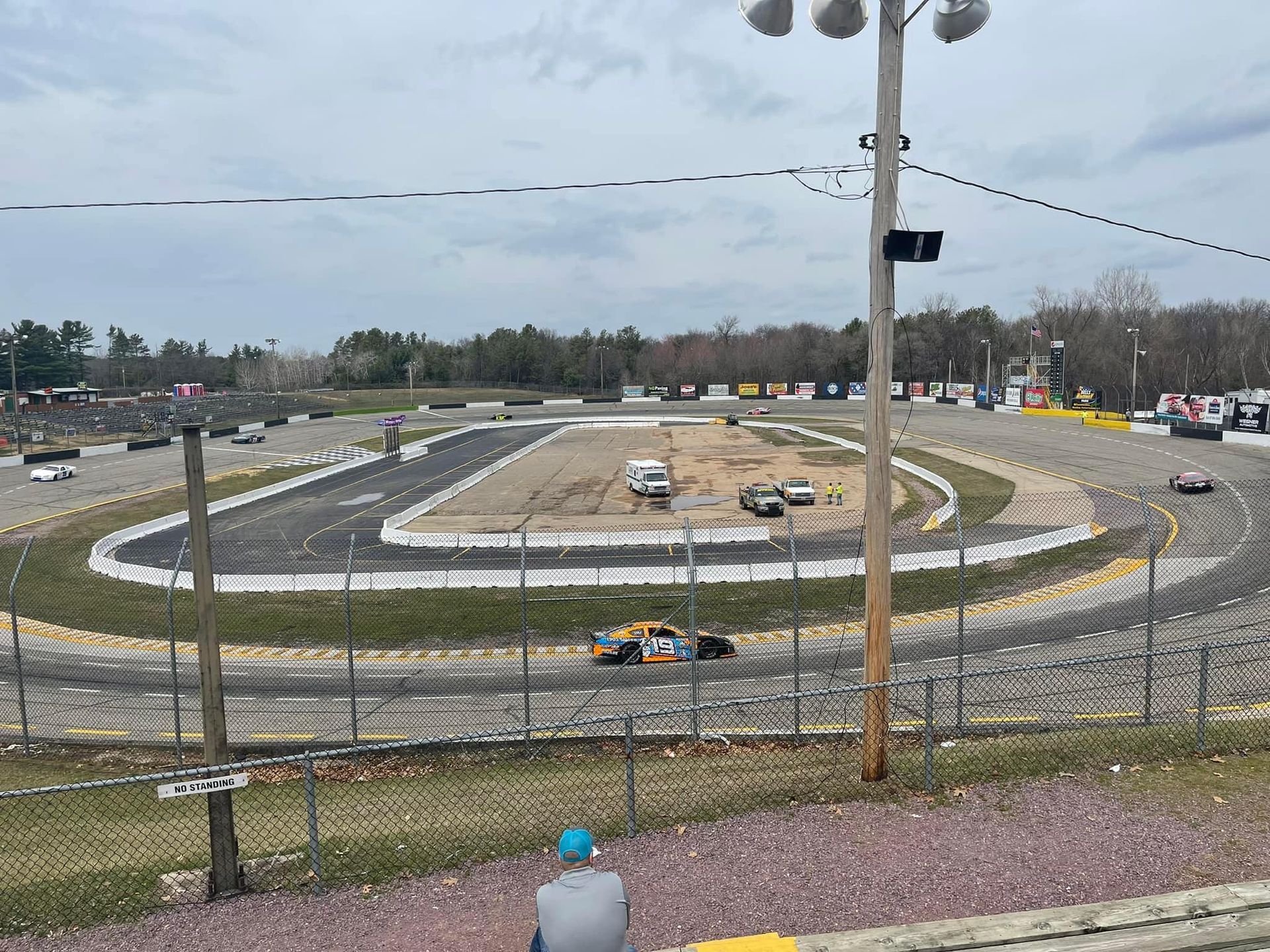 A race track with cars competing; view from the stands.  Green track, yellow barriers, overcast sky.