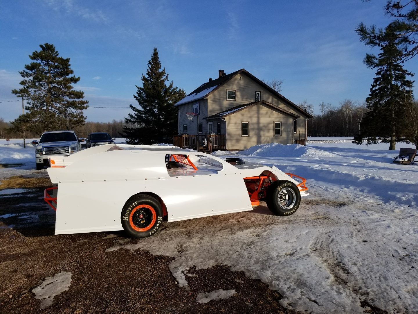White race car parked in a snowy yard in front of a house on a sunny day.