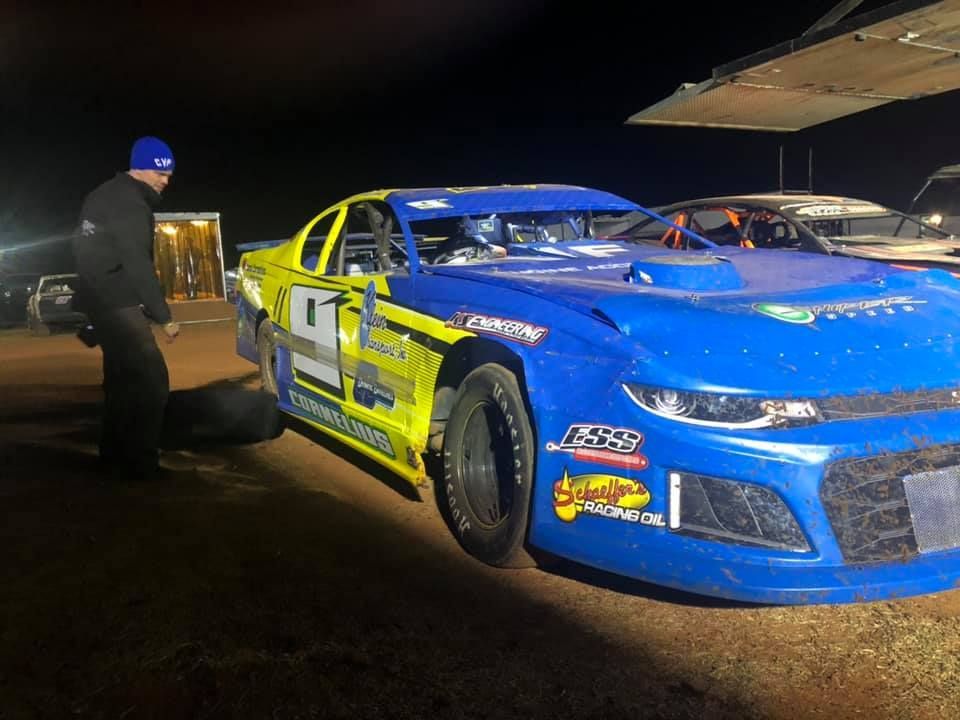 White race car parked in snow, in front of a house on a sunny day. The car has orange accents on the wheels and frame.