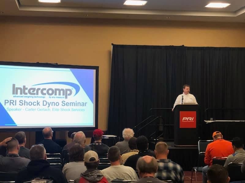 A man lecturing at a PRI Shock Dyno Seminar by Intercomp. A crowd of people sits in front of the presenter in a conference room.