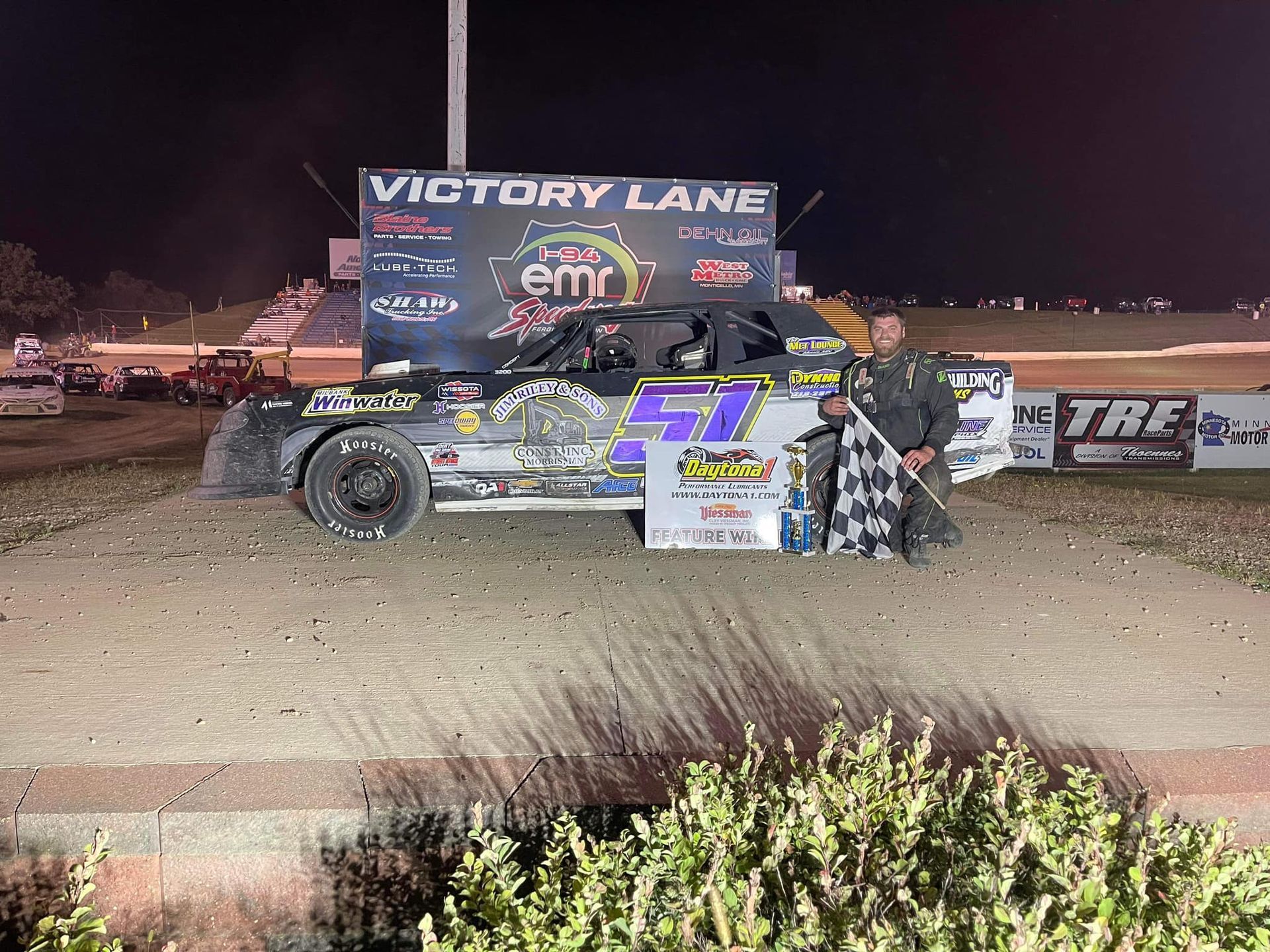 Race car driver holding checkered flag in victory lane after winning a race. Black car with number 57, night setting.