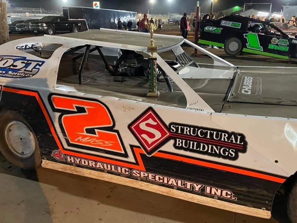 Race car with the number 2, a trophy, and other race cars at a dirt track at night. Sponsors' logos visible.