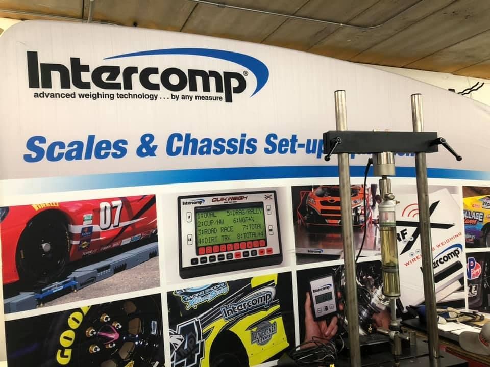 Intercomp booth with racing scales and chassis setup equipment. A digital scale displays readings; a racing car image is in the background.