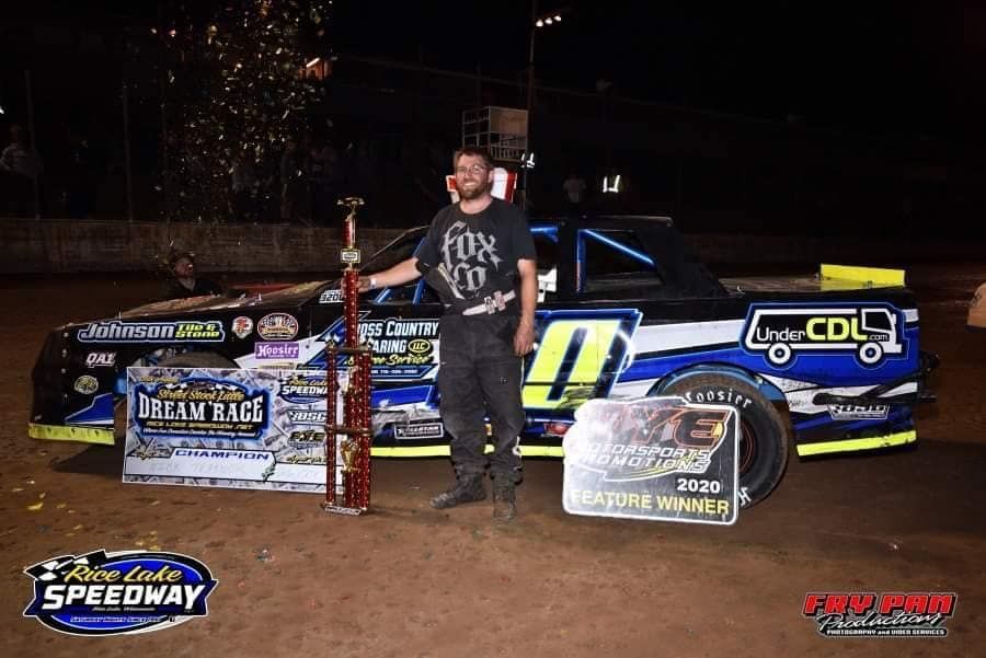 Race car driver stands with a trophy and car after a win at the Macon Speedway. The car is black, blue, and white.