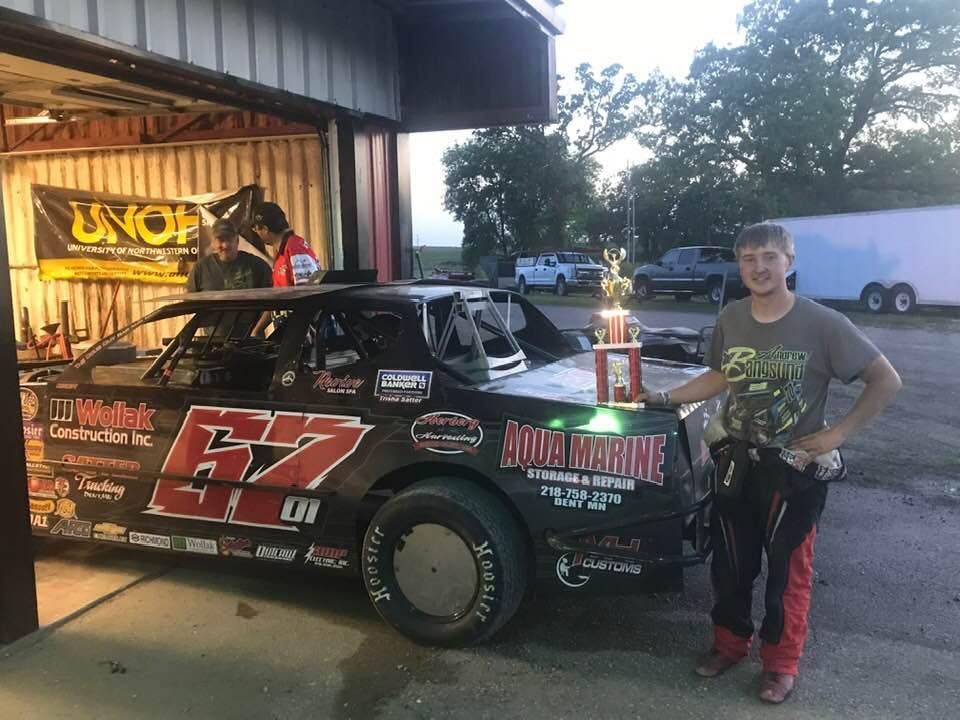 A young man in racing gear poses with a trophy next to his black race car, #67, in a garage setting.