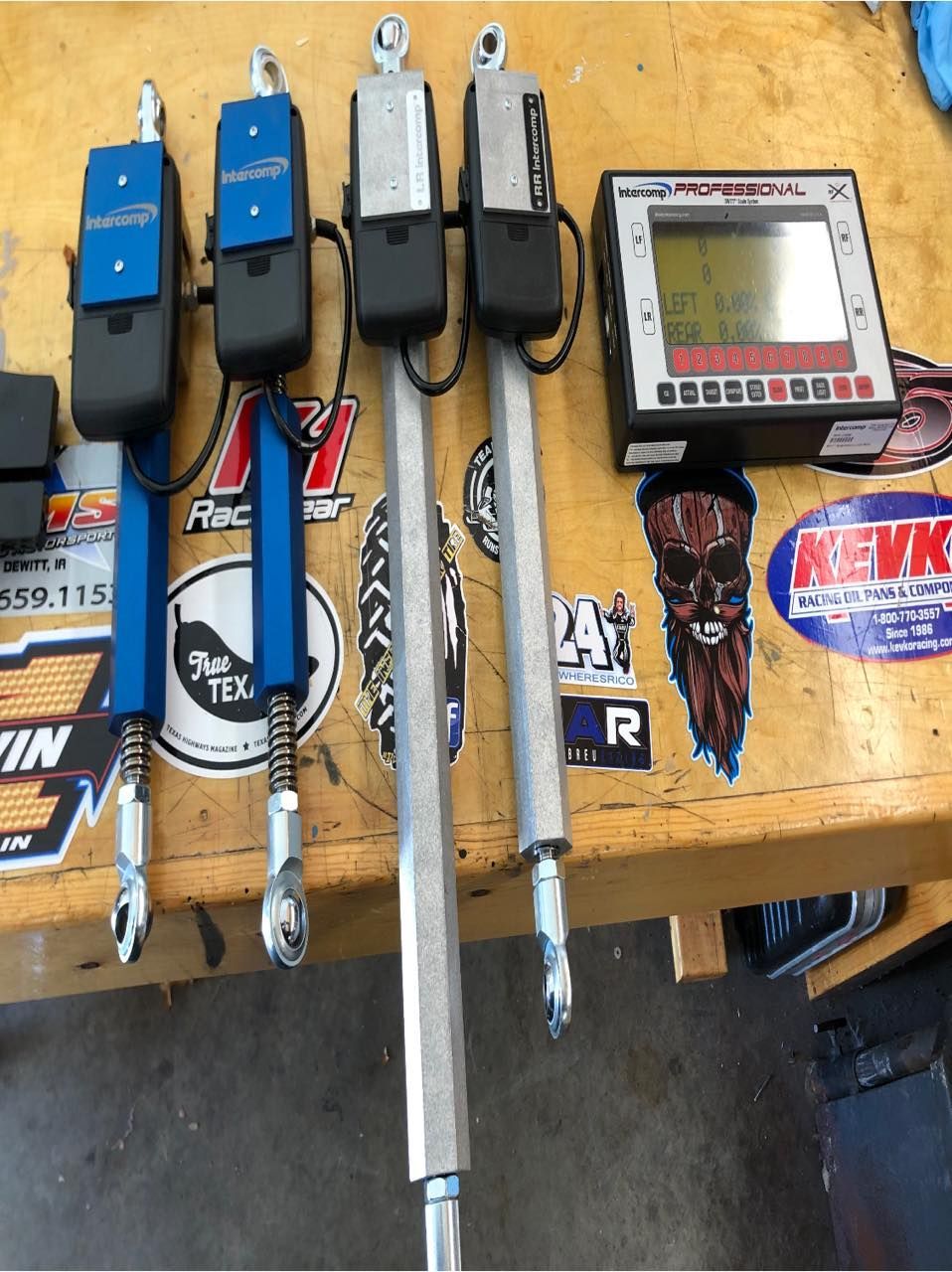 Four suspension scales and a digital display unit on a workbench, likely for vehicle setup adjustments. Blue and silver components are visible.